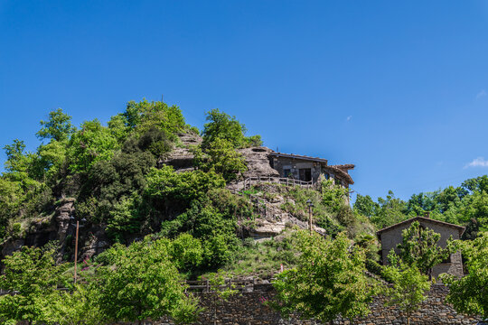 Stone house perched on rocky hillside in Rupit village, Catalonia, Spain, surrounded by lush greenery under clear blue sky