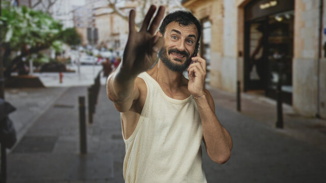 Man holding phone with raised hand waving and smiling on street amid buildings and pedestrians, wearing tanktop and beard; friendly greeting joy.