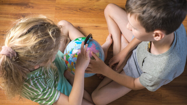 Two curious children sitting on wooden floor and looking at desk globe top view. Boy and girl studying geography pointing finger at map indoors. Early childhood education, homeschooling and travel pla