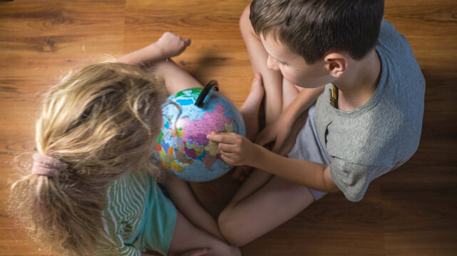 Two curious children sitting on wooden floor and looking at desk globe top view. Boy and girl studying geography pointing finger at map indoors. Early childhood education, homeschooling and travel pla