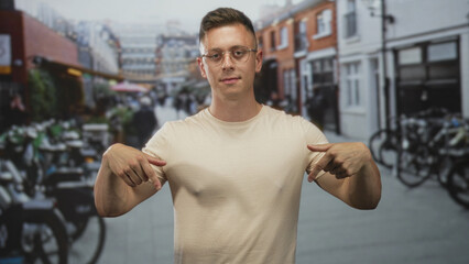 Fototapeta premium Man young blond handsome wearing beige t shirt points both index fingers at his chest and looks into camera on street lined with bicycles and shops; confidence.