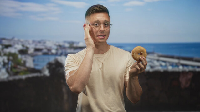 Man holding donut and touching cheek in a marina by the pier and sea, wearing glasses and a casual tshirt; hesitation hunger.