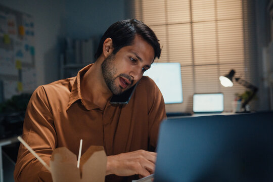 Young Latin businessman taking urgent phone call while reading accounting document on laptop at desk in dark night office. Multitasking overtime, overworked deadline, tired paperwork rush concept.