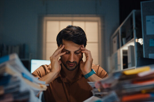Stressed young Latin businessman having severe headache and migraine at desk in night office. Debt crisis, corporate burnout, late overtime workload, mental health anxiety, business failure concept.