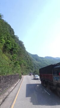 Vertical footage of a scenic paved highway winding through lush green mountain terrain in Nepal. The road passes between densely vegetated hills with utility lines and roadside safety barriers