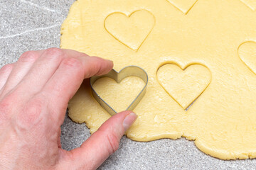 Obraz premium Pastry chef's hand cuts heart-shaped cookies from raw yellow dough on a gray table background, close-up.