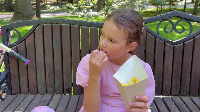Young European girl holds a brown paper container and is seen eating french fries, taking bites and chewing