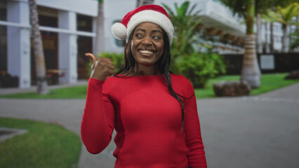 Fototapeta premium Woman wearing santa hat and red sweater gives thumbs up while smiling in front of building; holiday joy.