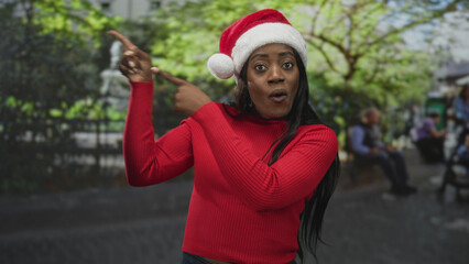 Fototapeta premium Woman in santa hat pointing finger to upper left, wearing red sweater on street near fountain and bench with animated expression; holiday joy.