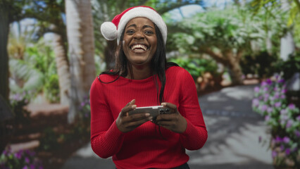 Fototapeta premium Woman wearing a santa hat holding a smartphone and laughing while playing a mobile game on a sunlit garden path with palms and flowers; holiday joy.