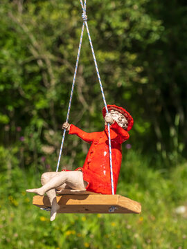 Red-Coated Ceramic Figurine On Wooden Swing In Sunny Garden Landscape With Playful Summer Mood