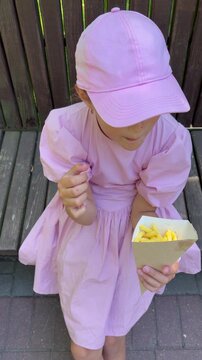 A 7-year-old girl in a pink dress eats French fries from a cardboard bag on a bench. Takeaway food