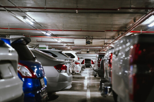 Rows of parked cars inside a modern underground parking garage with concrete ceiling, fluorescent lights, and red fire protection pipes.