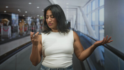 Fototapeta premium Woman holding smartphone to ear with hand on airport moving walkway, head tilted with weary expression and closed eyes while standing near gate; bored waiting.
