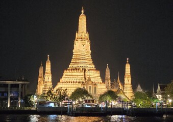 Naklejka premium Illuminated Wat Arun temple with golden spires reflecting on the Chao Phraya River at night in Bangkok, Thailand