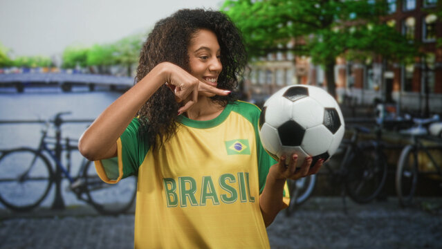 Teenage woman with curly hair wearing brasil t shirt holds soccer ball in hand and makes a celebratory fist gesture on street; team pride.