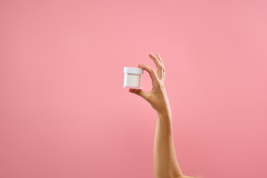 Hand holding small white cosmetic cream jar on pink copy space background. Concept of compact cosmetic packaging, skincare promotion, and facial moisturizer ads.