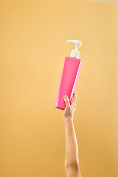 Hand holding bright pink pump bottle with skincare product on soft yellow background. Concept of daily skincare routine, and body care advertising, and colorful beauty branding.