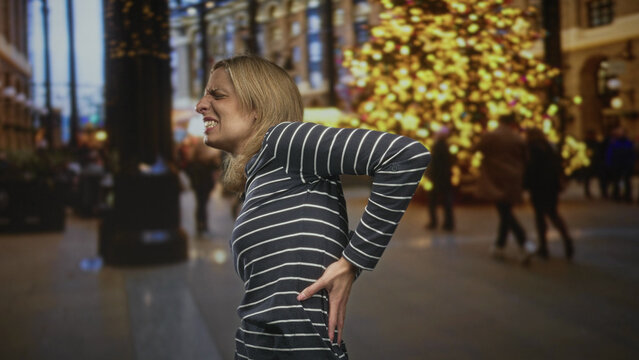 Woman holding hand to lower back and grimacing amid festive tree and blurred crowd in a building; pain backache.