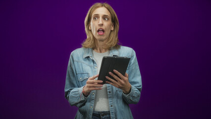 Woman holds tablet and taps touchscreen with worried gaze in studio purple backdrop  surprise reaction. © Krakenimages.com