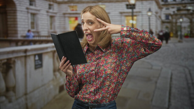 Blonde woman reading from a black paperback book and flashing a v sign near her eye while standing on a cobbled city street; playful surprise.