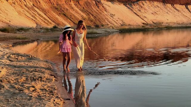 Mother and daughter walk barefoot along a lakeshore at sunset, peaceful family bonding, summer evening, maternal love, outdoor lifestyle.