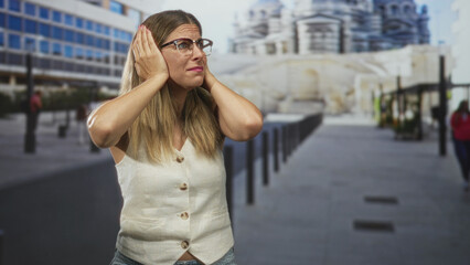 Fototapeta premium Woman covers ears with hands and glasses on street near monument and bollards, wearing sleeveless vest and jeans; stress discomfort.