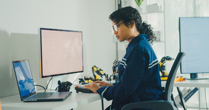 Young African American female robotics engineer coding AI software for robotic arm in laboratory. Embedded system, firmware testing, machine learning, industrial automation, future technology concept.