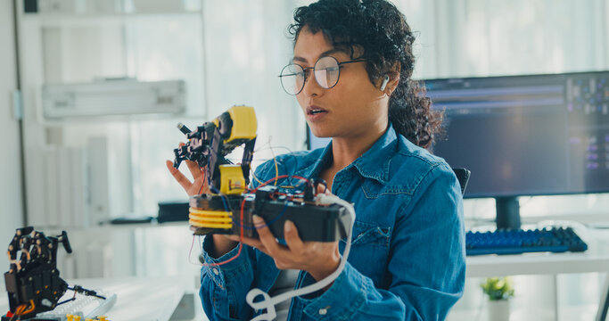 Young African American female robotics engineer discussing mechanical part on video call and looking at camera in laboratory. Remote work, virtual training, online meeting, future technology concept.