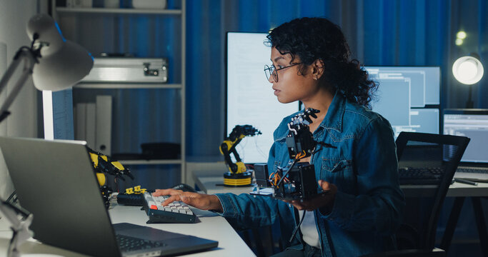 Young African American female programmer coding AI software on multiple screens in night office. Network security, cybersecurity, machine learning, working late, overtime, robotics automation concept.