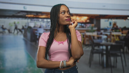 Fototapeta premium Black woman with hand on forehead and closed eyes, arms crossed in airport food court wearing pink shirt; travel delay frustration.