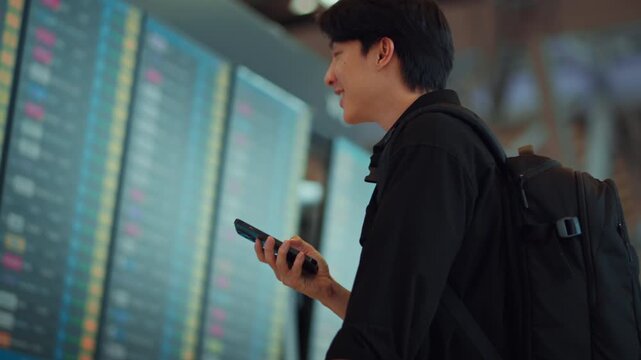 Young Asian man using smartphone while checking flight information board at airport terminal, digital travel and passenger journey concept