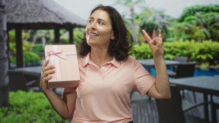 Fototapeta premium Woman holds pink gift box and makes rock horns gesture while smiling in a studio; playful celebration joy.