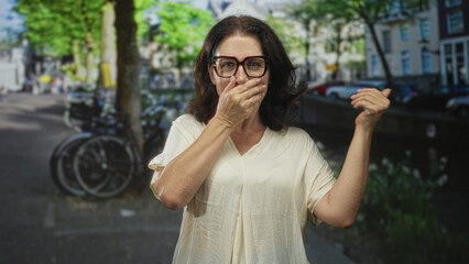 Fototapeta premium Woman covers mouth with hand and points thumb over shoulder on street by a canal wearing glasses and cream blouse; surprise caution.