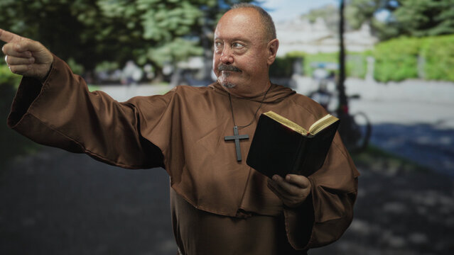 Senior hispanic man monk holding bible and cross necklace points finger forward on street path near park; devotion faith guidance.