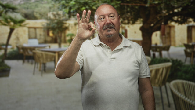 Man makes ok sign with raised right hand, visible mustache and white polo in building patio cafe setting; approval contentment.