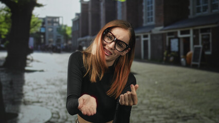 Fototapeta premium Redhead woman in black long sleeve top and glasses extends outstretched palm asking on a cobblestone street near brick storefronts; pleading request.