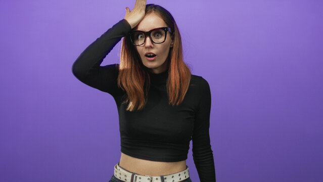 Woman redhead wearing glasses places hand on forehead against purple studio backdrop; playful forgetfulness.