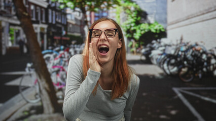 Fototapeta premium Woman cupping hand to mouth shouting on a city street with parked bicycles and buildings visible; joy celebration.