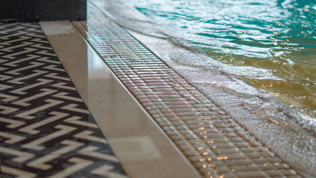 Water gently laps against the tiled overflow gutter and elegant patterned floor surrounding a swimming pool