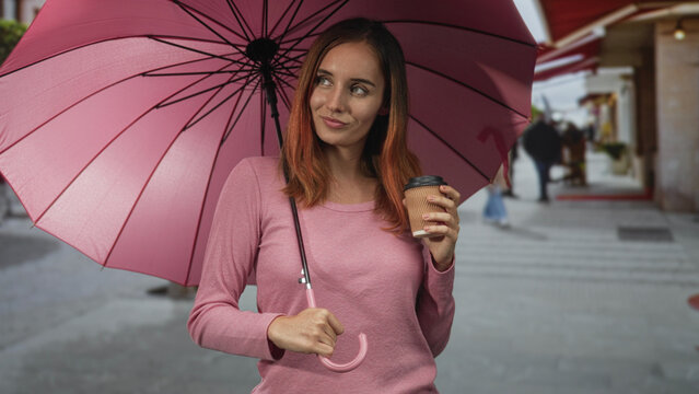 Woman redhead holding pink umbrella and takeaway coffee cup, right hand gripping handle, slight smile and relaxed posture on a street lined with shops; contentment.