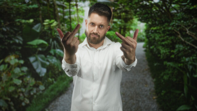 Man with beard in white shirt shows both middle fingers toward camera on a leafy forest path, hands extended and forward; defiance.