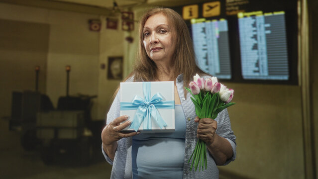 Senior hispanic woman holds blue ribbon gift box and pink tulip bouquet while smiling in an airport check in area by the departure display; joy reunion celebration.