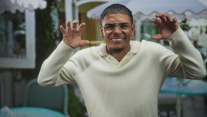 Fototapeta premium Young hispanic man in cream sweater wearing glasses, smiling and raising hands in a playful claw gesture at a street cafe; playful.
