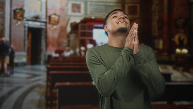 Man with hands clasped in prayer inside ornate church nave, smiling gently in green shirt; reverence faith.