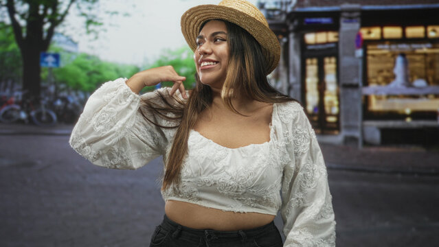Woman wearing straw hat touches hair with hand near head, white embroidered crop top showing midriff on a street lined with shops; playful confidence.