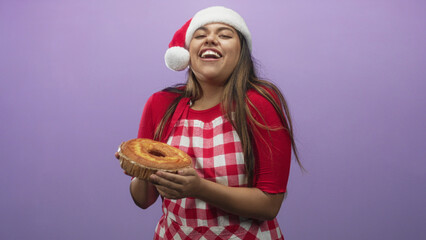 Fototapeta premium Plus size woman holding sponge cake with hands, smiling, wearing santa hat and checkered apron in studio with purple backdrop; holiday joy.
