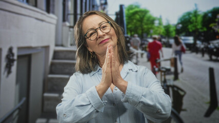 Fototapeta premium Woman with hands pressed palms together in prayer on street beside building stairs, wearing glasses and light blue shirt; serenity.