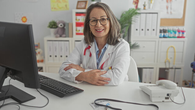 Hispanic senior woman doctor seated at desk smiling with folded arms and visible stethoscope, computer keyboard and pediatric toys in clinic; calm caring professionalism.