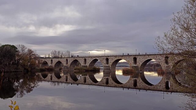 Masonry Pointed arch Stone bridge (Puente de Piedra) across the river Duero in Zamora, Castile-Leon, Spain. Reflection in the water. Autumn nature. Pedestrians walking on the bridge. Overcast.